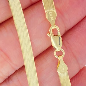 Könnte beinhalten: Eine goldfarbene Fischgräten-Kettenhalskette mit einem Karabinerverschluss. Die Halskette ist flach und flexibel, mit einer glatten, reflektierenden Oberfläche. Der Verschluss ist mit "14 KT" markiert.