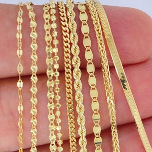 May include: A collection of delicate gold necklaces in various chain styles. The chains include a variety of designs, such as oval links, beaded, rope, and herringbone patterns. The necklaces are displayed against a neutral background, highlighting their shiny, golden appearance.