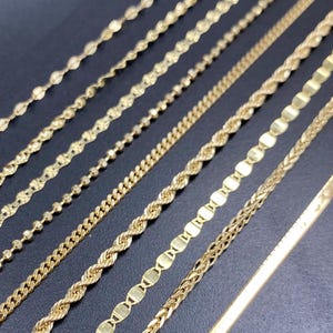 May include: A collection of gold necklaces displayed on a dark surface. The necklaces vary in style, showcasing different chain designs such as beaded, rope, curb, and flat styles. The chains are all a shiny gold color, reflecting light.