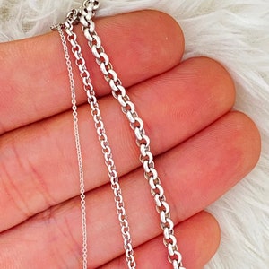 Puede incluir: Primer plano de tres collares plateados con diferentes estilos de cadena. Las cadenas se muestran sobre un fondo blanco y suave. Los collares son delicados y parecen estar hechos de plata de ley.
