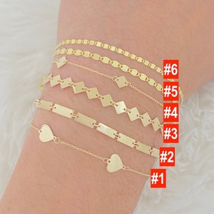 K&ouml;nnte beinhalten: Sechs goldene Armb&auml;nder am Handgelenk einer Person. Die Armb&auml;nder sind von 1 bis 6 nummeriert. Die Armb&auml;nder sind aus 14 Karat Gold gefertigt und haben verschiedene Designs, darunter ein Herz, eine Kette und ein quadratisches Muster.