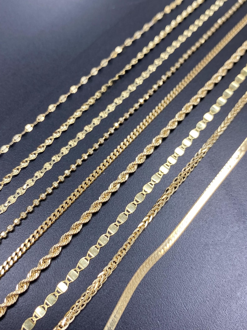 May include: A collection of gold necklaces displayed on a dark surface. The necklaces vary in style, showcasing different chain designs such as beaded, rope, curb, and flat styles. The chains are all a shiny gold color, reflecting light.