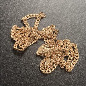 Pu&ograve; includere: Una collana a catena color oro con un design a maglie a catena. La collana &egrave; appoggiata su una superficie scura. La catena &egrave; in metallo e ha una finitura lucida. La chiusura &egrave; visibile.