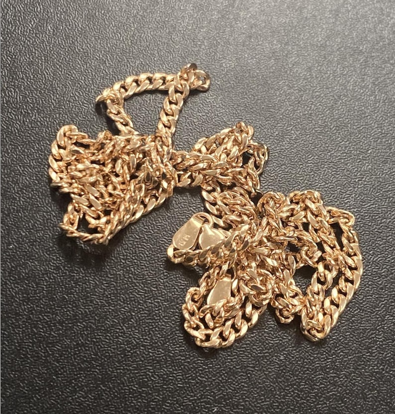 Pu&ograve; includere: Una collana a catena color oro con un design a maglie a catena. La collana &egrave; appoggiata su una superficie scura. La catena &egrave; in metallo e ha una finitura lucida. La chiusura &egrave; visibile.