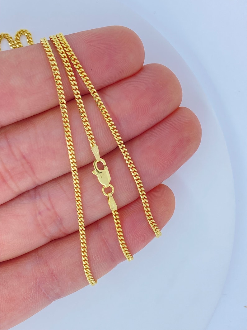 May include: A gold chain necklace with a curb link style. The chain is shown on a hand, with the clasp visible.