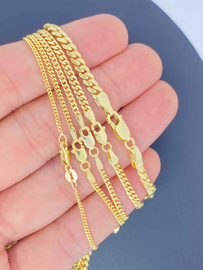 May include: Five gold chains of different widths and lengths, displayed on a hand.