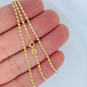 May include: A delicate gold necklace with a unique chain design. The chain features small, diamond-shaped links connected in a repeating pattern. The necklace is shown against a neutral background, highlighting its bright, polished finish. The clasp is visible, indicating the necklace is ready to wear.