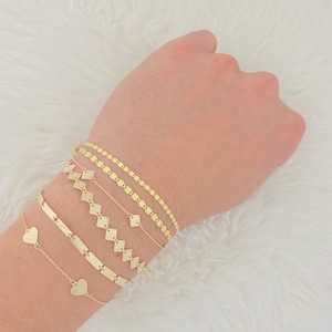 K&ouml;nnte beinhalten: Ein Set aus f&uuml;nf goldenen Armb&auml;ndern, darunter eine zarte Kette mit Herzanh&auml;ngern, ein schlichtes Bar-Armband, eine Kette mit quadratischen Gliedern und zwei Ketten mit rautenf&ouml;rmigen Gliedern.