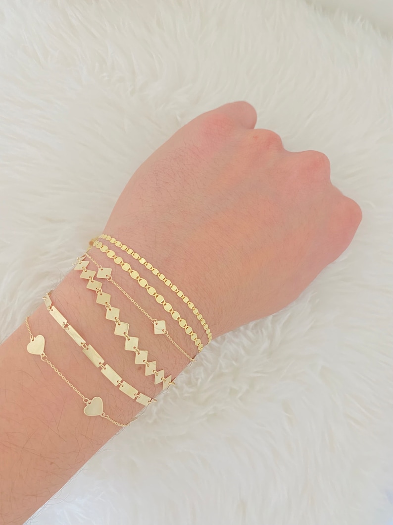K&ouml;nnte beinhalten: Ein Set aus f&uuml;nf goldenen Armb&auml;ndern, darunter eine zarte Kette mit Herzanh&auml;ngern, ein schlichtes Bar-Armband, eine Kette mit quadratischen Gliedern und zwei Ketten mit rautenf&ouml;rmigen Gliedern.