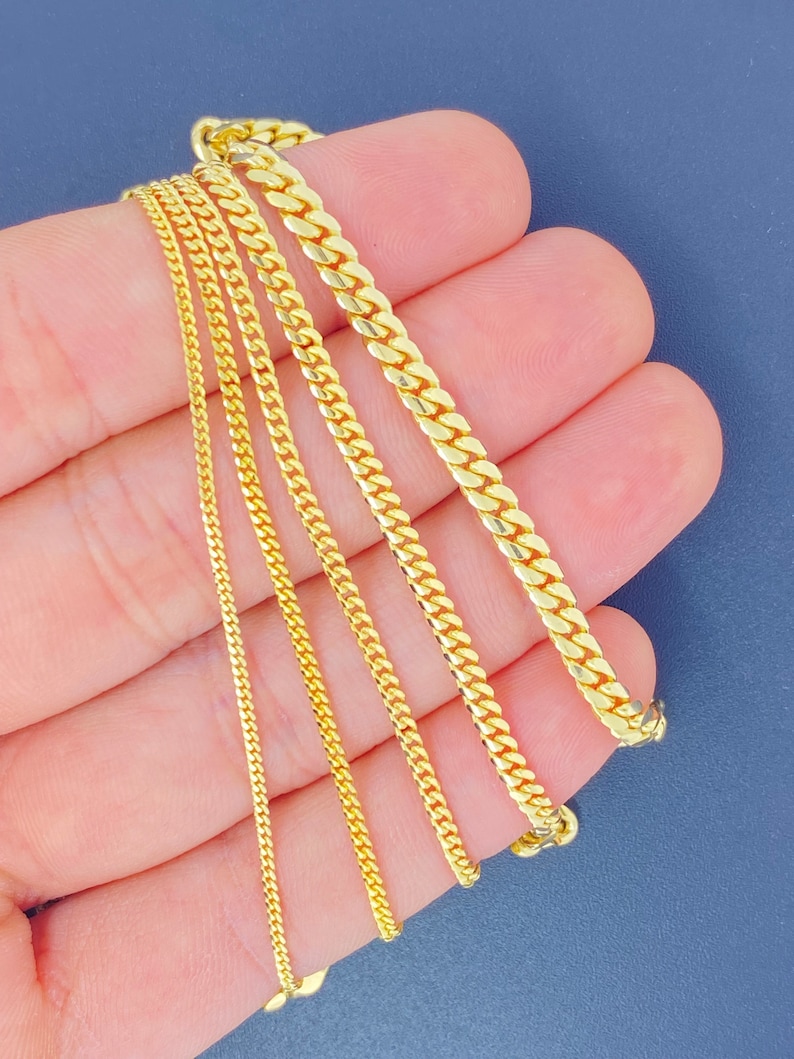 May include: Five gold chains of different widths and lengths, displayed on a hand.