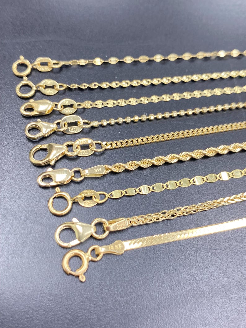 May include: A collection of gold necklaces with various chain styles, including beaded, rope, and flat designs. Each necklace features a clasp for secure wear. The chains are displayed against a dark background.