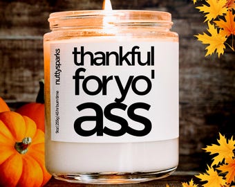 May include: A lit candle in a glass jar with the text "thankful for yo' ass" on a white label. The candle is 255g and has a 45-hour burn time. The background includes pumpkins and fall leaves.