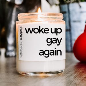 May include: A lit candle in a clear glass jar with a white label. The label reads "woke up gay again" in black text. The jar is labeled "nutty sparks" and indicates a 9oz (255g) size with a 45-hour burn time.