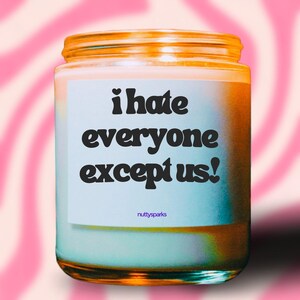 May include: A glass jar candle with light blue wax and a gold lid. The label displays the text "i hate everyone except us!" in black. The background features a pink and white wavy pattern.