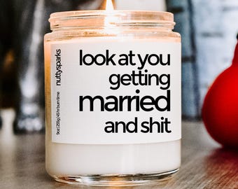 Puede incluir: Una vela encendida en un tarro de cristal con el texto "look at you getting married and shit" en negro. La vela está etiquetada como "nutty sparks" y tiene un tamaño de 255g con un tiempo de combustión de 45 horas.