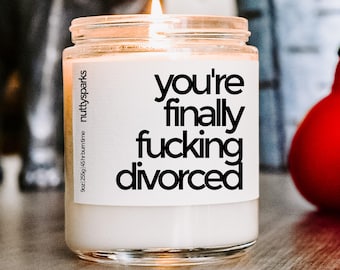 Könnte beinhalten: Eine brennende weiße Kerze in einem Glas mit dem Text "you're finally fucking divorced" in Schwarz. Das Etikett enthält auch "nutty sparks" und "9oz | 255g | 45 hr burn time".