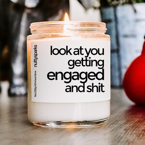 Op de afbeelding: Een brandende witte kaars in een heldere glazen pot. De pot heeft een label met de tekst "look at you getting engaged and shit". De kaars is van "nutty sparks" en heeft een brandtijd van 45 uur.