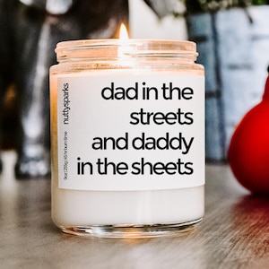May include: A lit candle in a clear glass jar with a white label. The label reads "dad in the streets and daddy in the sheets". The jar is 9 oz (255g) and has a 45-hour burn time.