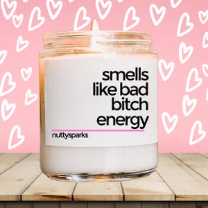 May include: A lit white candle in a glass jar with the text "smells like bad bitch energy" on a white label. The brand name "nuttysparks" is at the bottom of the label. The background is pink with white heart shapes.