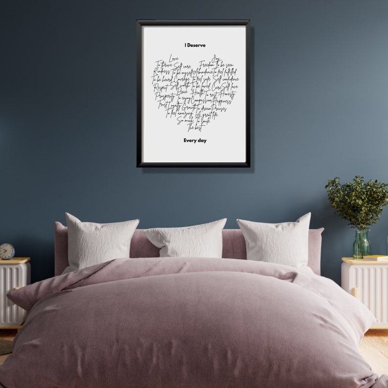 K&ouml;nnte beinhalten: Ein schwarzer gerahmter Druck mit wei&szlig;em Hintergrund und schwarzem Text, der "Ich verdiene" lautet und eine Liste positiver Affirmationen enth&auml;lt. Der Text ist handschriftlich und in Herzform angeordnet. Der Druck h&auml;ngt an einer blauen Wand &uuml;ber einem Bett mit einer rosa Bettdecke und wei&szlig;en Kissen.