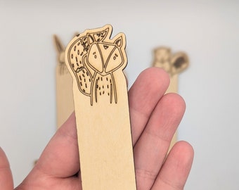 May include: A wooden bookmark with a laser-cut design of a fox with a bushy tail and leaves at the bottom. The bookmark is light brown and has a blank space for writing.