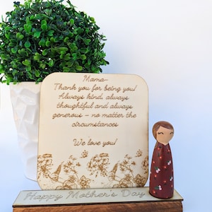 May include: A wooden Mother's Day gift set featuring a decorative wooden sign with the text "Mama" and a heartfelt message, along with a wooden peg doll. The base reads "Happy Mother's Day". A green plant in a white pot is in the background.
