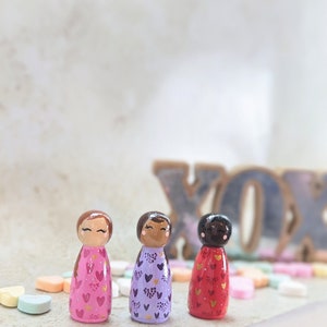 May include: Three wooden peg dolls with painted faces and hearts on their dresses. The dolls are wearing pink, purple, and red dresses with gold hearts. The dolls are standing on a white surface with candy hearts scattered around them.