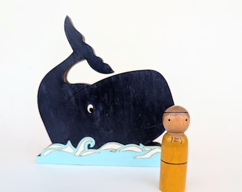 May include: A wooden toy whale with a black silhouette and a blue and white ocean base. A wooden peg doll with a brown head and a yellow shirt stands next to the whale.