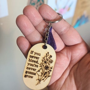 May include: Wooden keychain with a purple tassel and the text "If you never bleed, you're never gonna grow" with a floral design.
