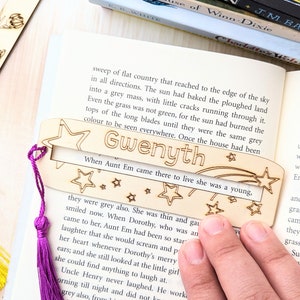 May include: A wooden bookmark with the name "Gwenyth" and star designs, accented with a purple tassel. The bookmark is placed on an open book, with other books stacked in the background.