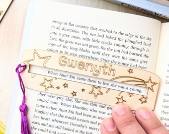 May include: A wooden bookmark with the name "Gwenyth" and star designs, accented with a purple tassel. The bookmark is placed on an open book, with other books stacked in the background.