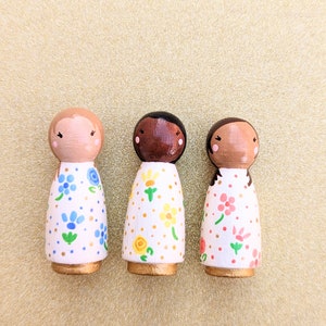 May include: Three wooden peg dolls with painted faces and floral patterned dresses. The dolls are standing in a row on a gold background. The dresses are white with different colored flowers.