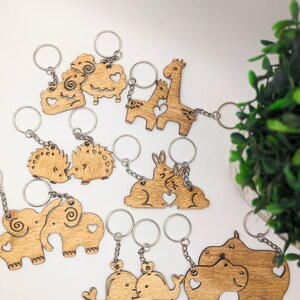 May include: A set of 10 wooden keychains featuring different animal couples, including elephants, hippos, whales, giraffes, hedgehogs, and sheep. Each keychain is made of natural wood and has a heart-shaped cutout in the center.
