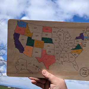 May include: A wooden map of the United States with each state outlined in a different color. The map is held in a hand against a blue sky with white clouds. The map is labeled with the state abbreviations. The map is made by Pepe and Data.