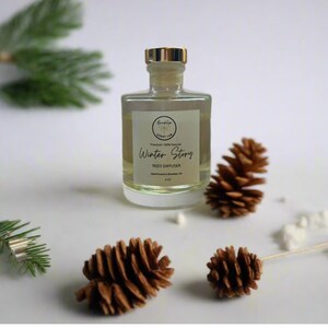 May include: A clear glass bottle with a gold lid filled with a reed diffuser. The bottle is labeled "Winter Story Reed Diffuser" and is sitting on a white surface with pine cones.