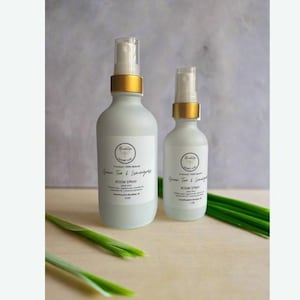 May include: Two glass bottles of room spray with gold caps and labels that say "Green Tea & Lemongrass Room Spray" in white text. The bottles are sitting on a light wood surface with green grass stems in the foreground.