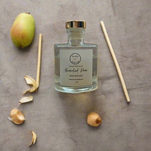 May include: A clear glass bottle with a gold lid and a white label that reads "Brandied Pear Reed Diffuser" on a light brown surface. The bottle is surrounded by pear peels and a wooden reed diffuser stick.