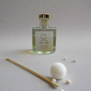 May include: A clear glass bottle with a gold lid and a white label that reads "Clean Cotton Reed Diffuser" on a light gray background. The bottle is surrounded by white decorative balls and a wooden reed diffuser stick.