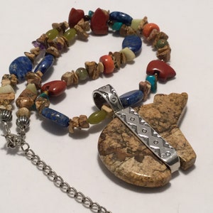 May include: A multi-coloured stone necklace with a large brown stone pendant shaped like a bear. The pendant has a silver band with a geometric pattern. The necklace has a silver chain.