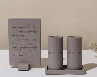 May include: A gray stone Sabbath candle holder set. The set includes two cylindrical candles with lit wicks, a rectangular base, and a rectangular plaque with Hebrew text. The candles and plaque are engraved with Hebrew text.