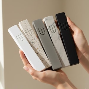 May include: Five rectangular mezuzah cases in varying colors and textures are held in a person's hands. The cases include white, speckled, gray, marble-patterned, and black. Each case has a recessed letter "U" on the front.