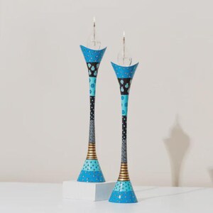 Op de afbeelding: Twee decoratieve kandelaars met brandende kaarsen. De houders zijn beschilderd met blauwe, zwarte en gouden patronen. De basis is lichtblauw met een stippenpatroon en de bovenkant is uitlopend met een vergelijkbaar ontwerp. Ongeveer 40 cm hoog.