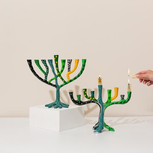 May include: Two green and blue menorahs with yellow and black accents. The menorahs are shaped like trees and have a total of nine candle holders. One menorah has a lit candle.