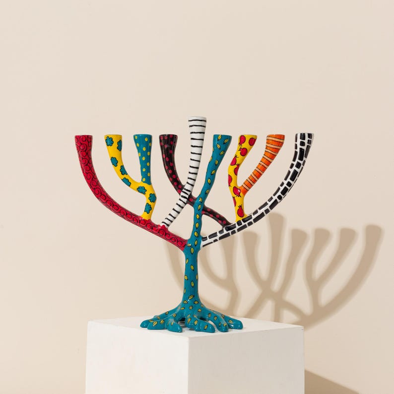 May include: A colorful menorah with a tree-like design. The menorah is painted in shades of red, yellow, blue, black, and white. The menorah is standing on a white pedestal.