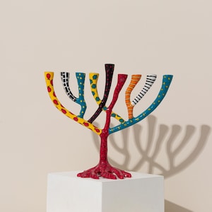 May include: A colorful menorah with a tree-like design. The branches are painted with different patterns, including red, yellow, blue, and black. The menorah is standing on a white surface.