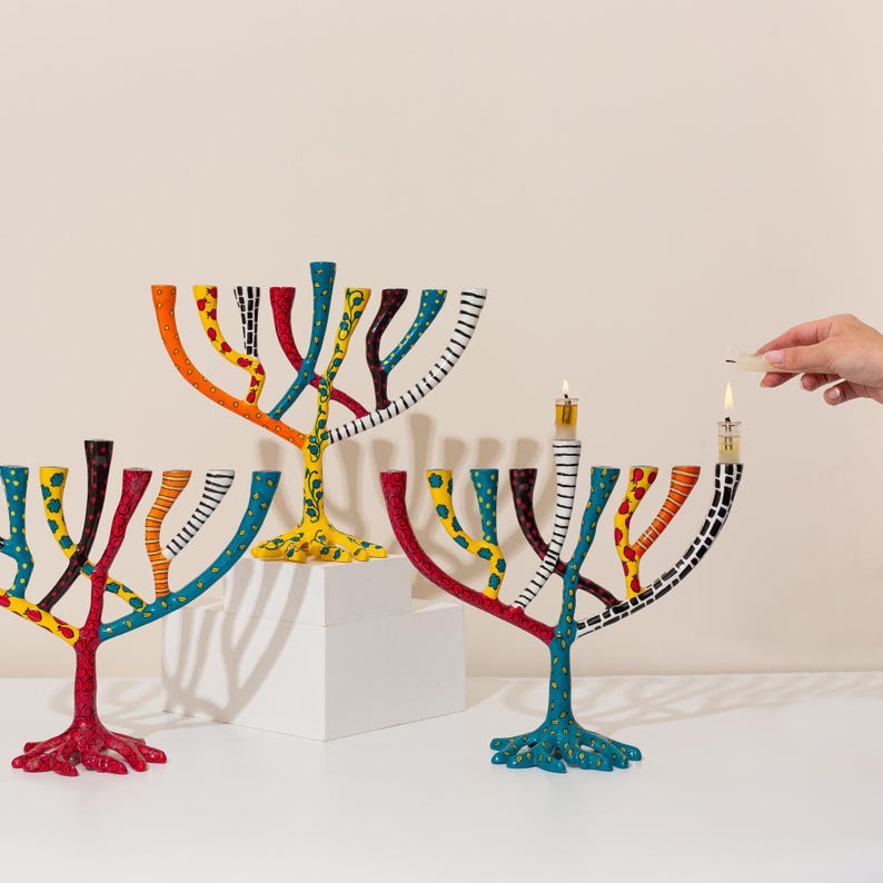 May include: Three colorful menorahs with a hand lighting a candle on one of them. The menorahs are made of metal and have a tree-like design with branches that hold the candles. Each menorah is painted in a different color scheme, with one being yellow, one being red, and one being blue.