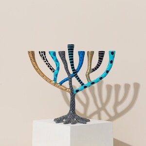 May include: A colorful menorah with a tree-like design. The menorah is painted in shades of blue, black, gold, and gray. The branches are decorated with different patterns, including polka dots, stripes, and leaves.