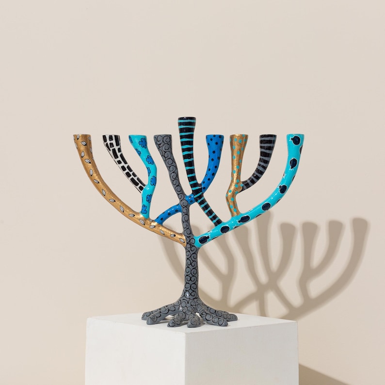 May include: A colorful menorah with a tree-like design. The menorah is painted in shades of blue, black, gold, and gray. The branches are decorated with different patterns, including polka dots, stripes, and leaves.