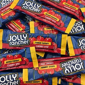 May include: A pile of Jolly Rancher Cherry Stix hard candy. The wrappers are blue with red and yellow accents, and the Jolly Rancher logo is prominently displayed. The candy sticks are red and feature a cherry design.