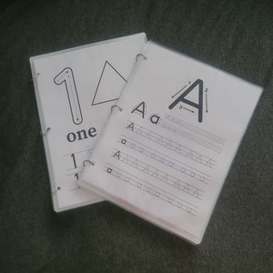 May include: Two white pages with clear plastic covers, bound together with silver rings. The left page shows the number "1" with arrows indicating the direction to trace the number. The right page shows the letter "A" with arrows indicating the direction to trace the letter. The page also has lines for practicing writing the letter "A".
